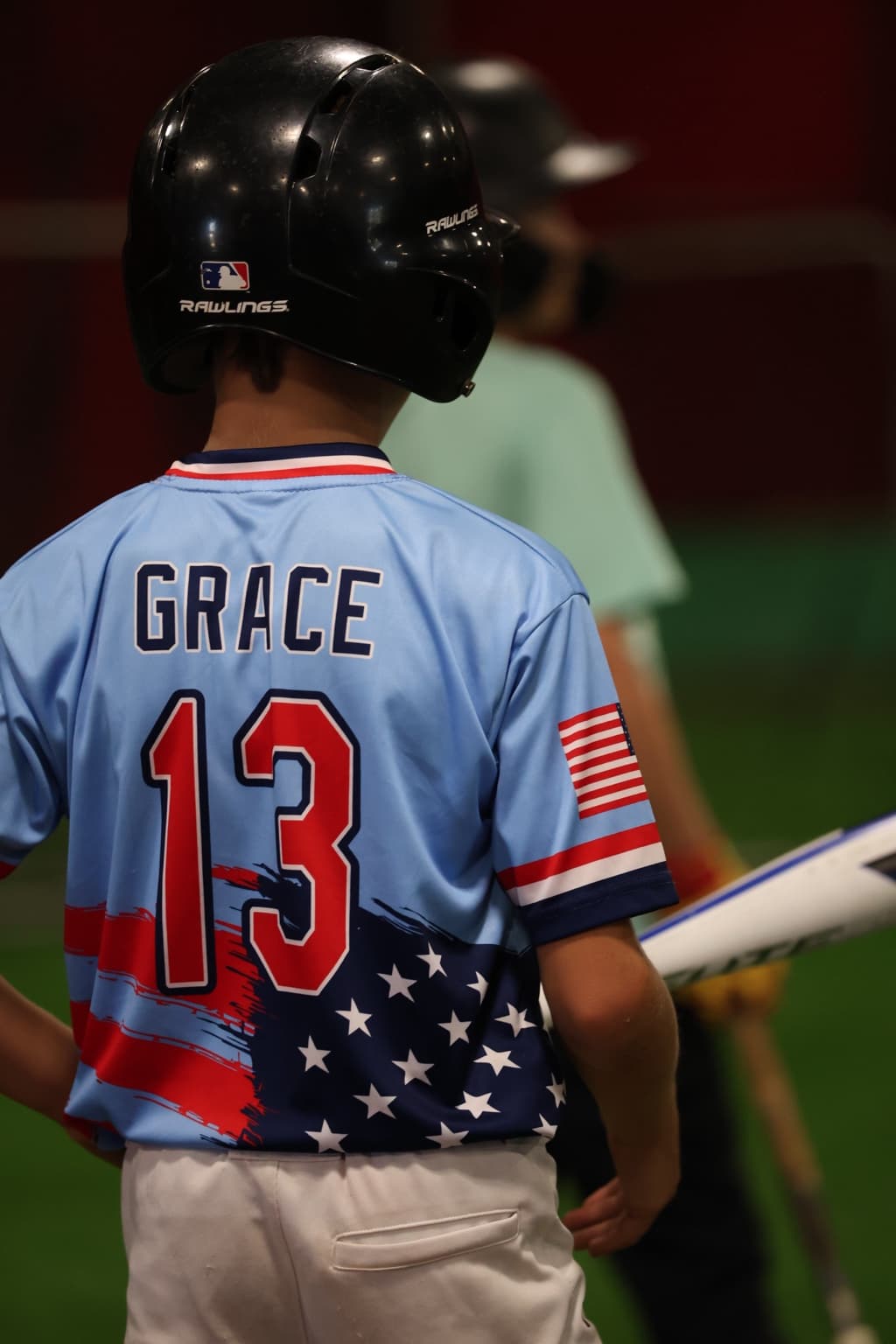 A Fall Camp player in a light blue Grace No. 13 jersey and batting helmet