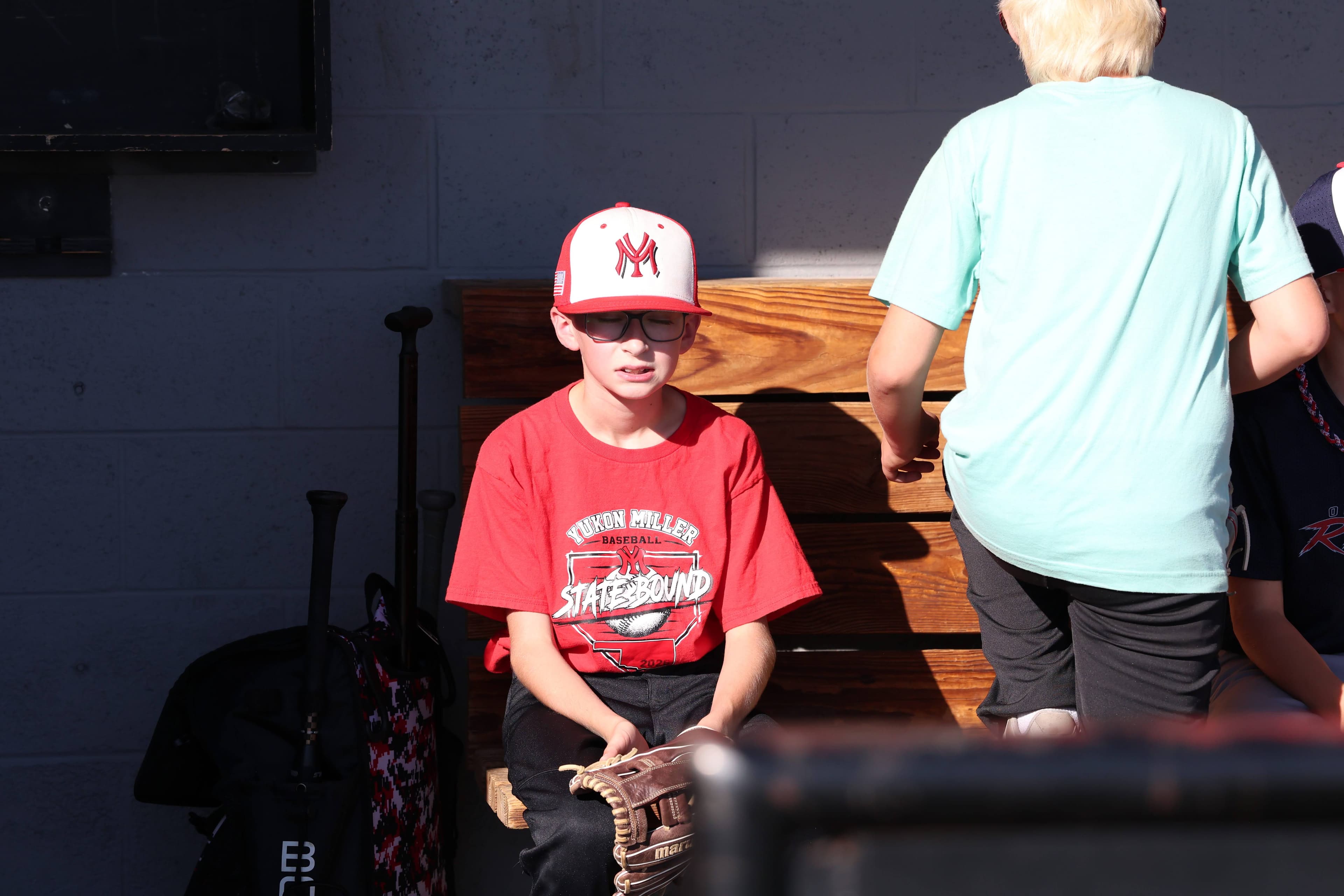A camper in a Yukon Miller Baseball shirt sits on the dugout bench