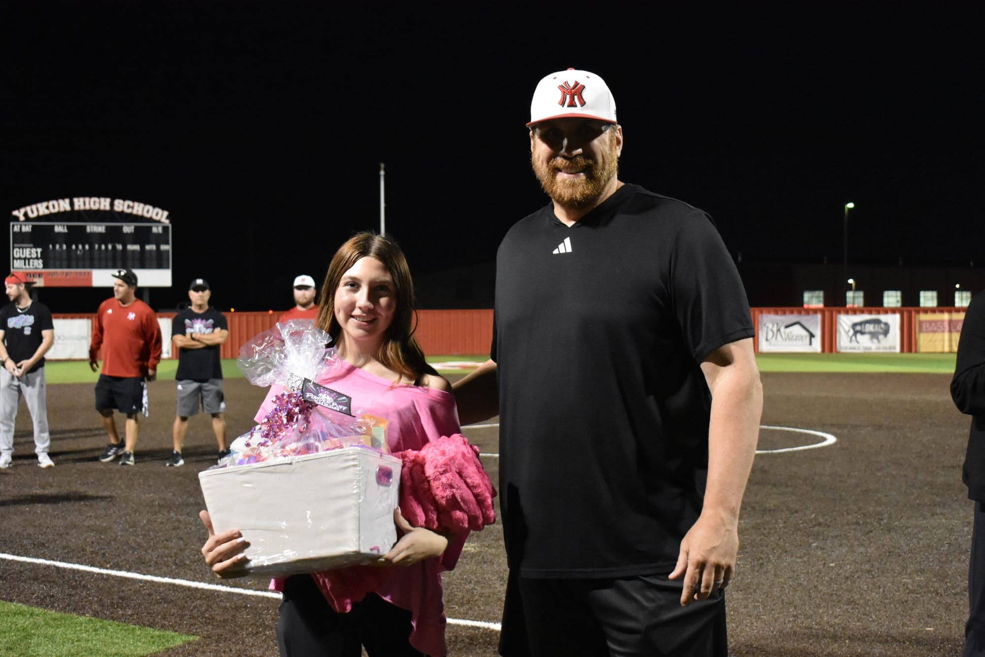A Powder Puff participant in a pink shirt holds a gift basket and a pink blanket beside an adult on the field