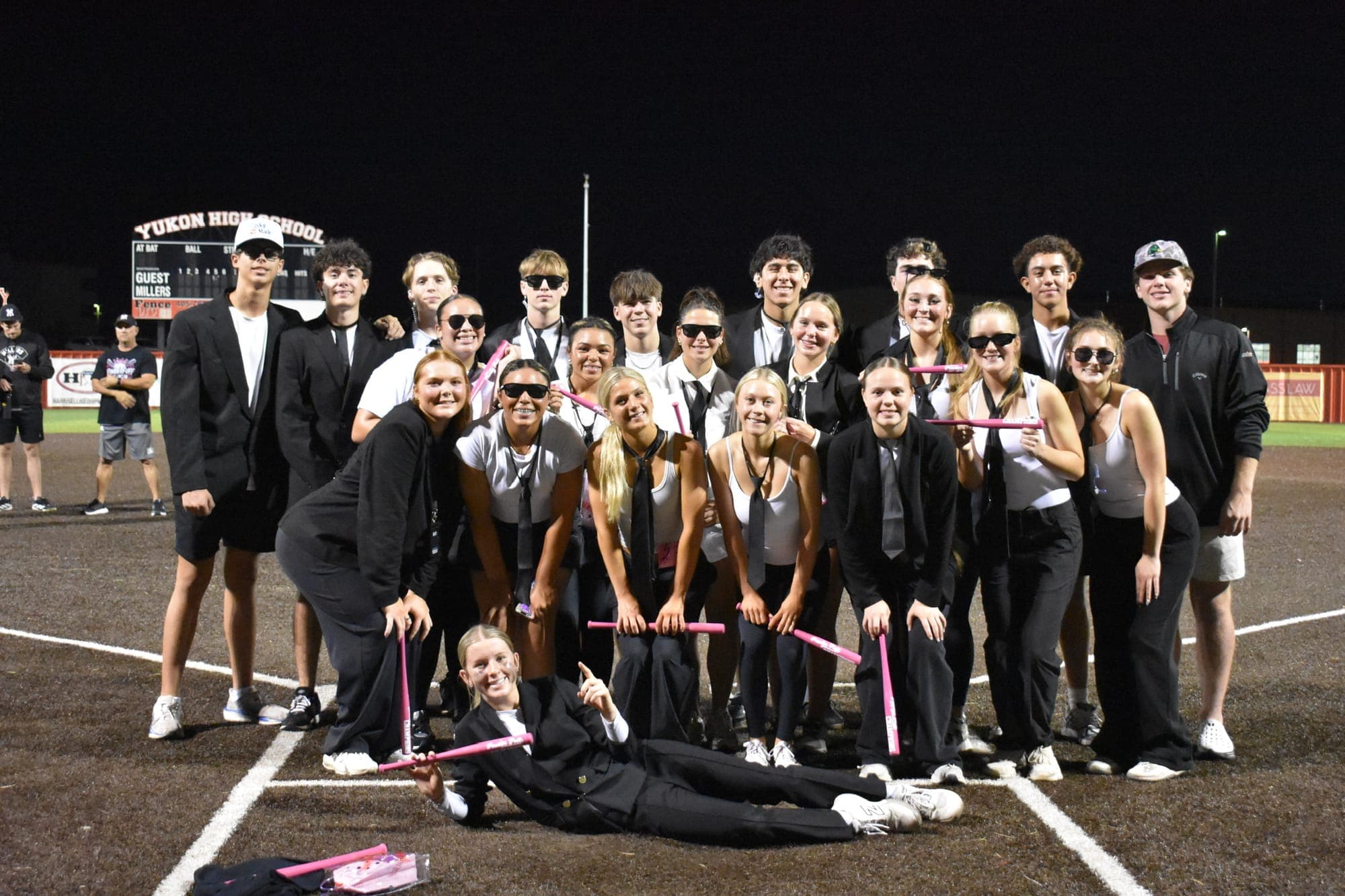 A Powder Puff team in black and white poses with pink bats in front of the Yukon High School scoreboard
