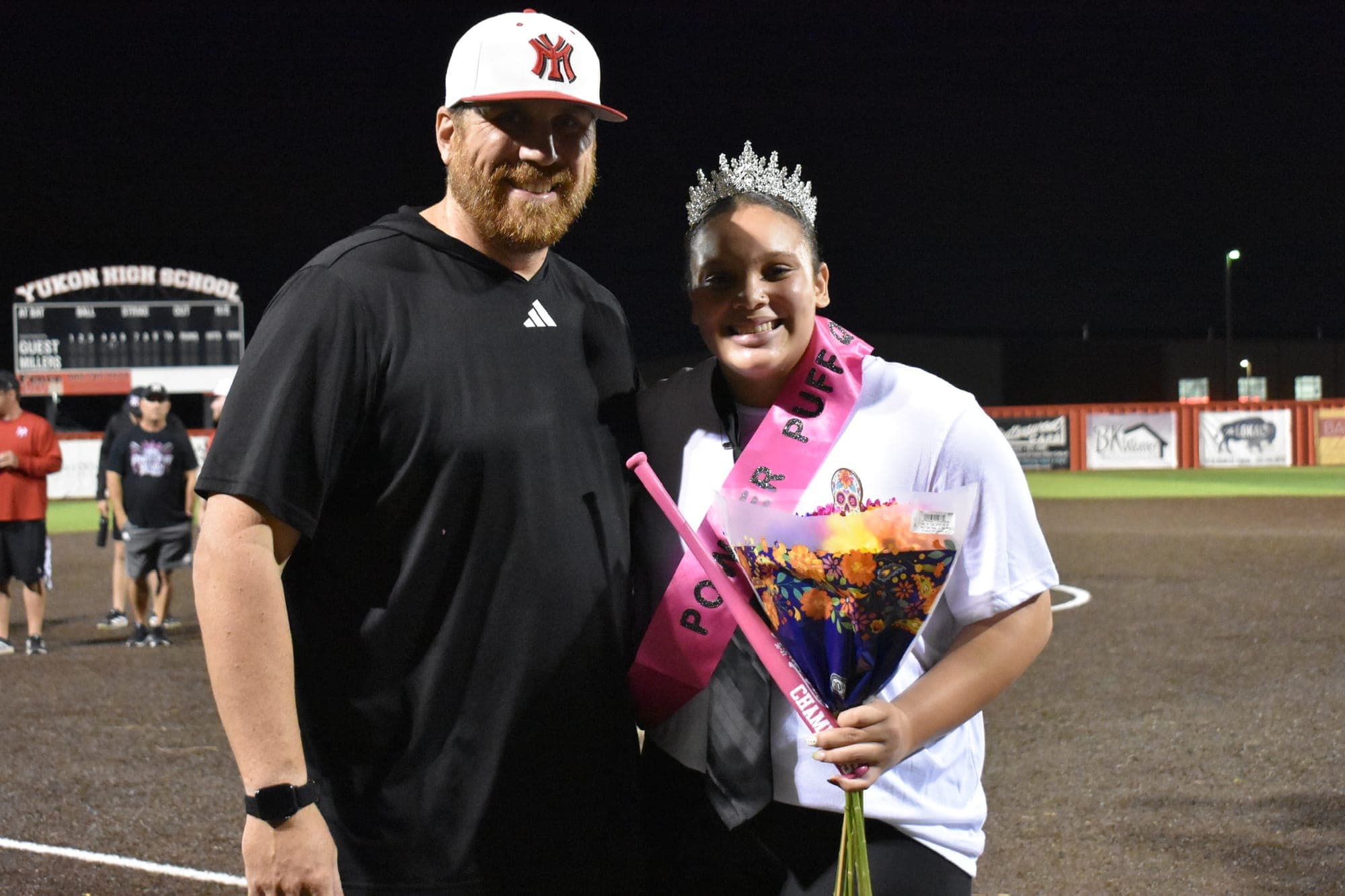 The Powder Puff queen, wearing a crown and sash, holds flowers and a pink bat on the field